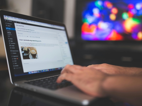 Hands typing on a laptop with a blog post visible, cozy indoor setting with colorful screen in background.